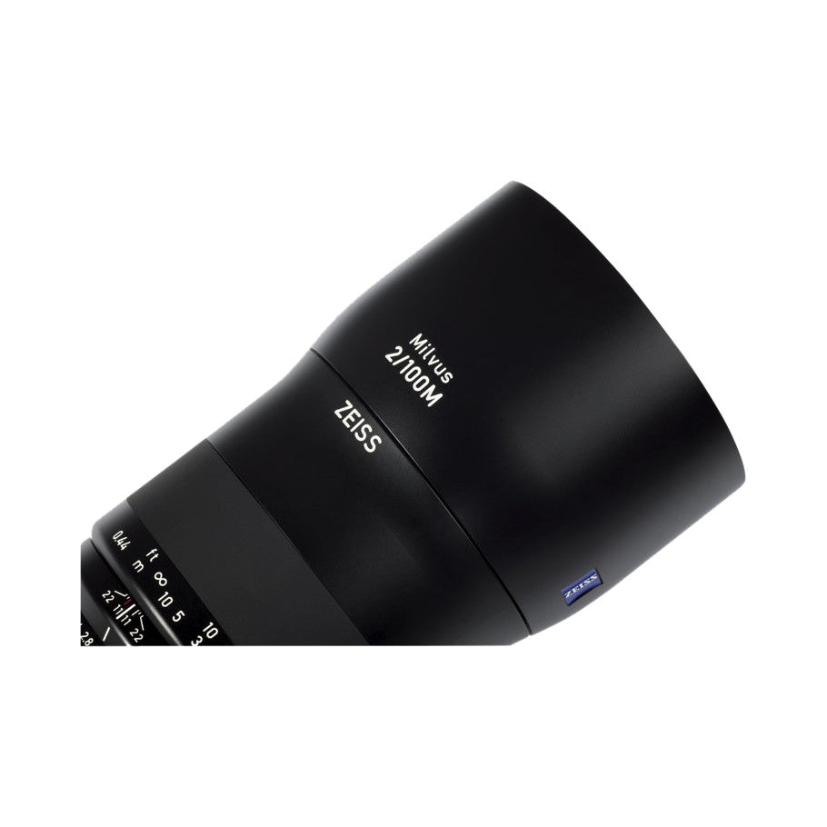 Close-up angled view of a black Zeiss Milvus 100mm f/2.0 macro lens showing barrel, focus ring, and distance scale markings.