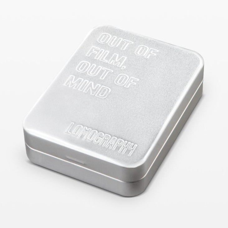Silver metal film case shown at an angle displaying the lid with embossed text reading "OUT OF FILM OUT OF MIND" and "LOMOGRAPHY" branding.