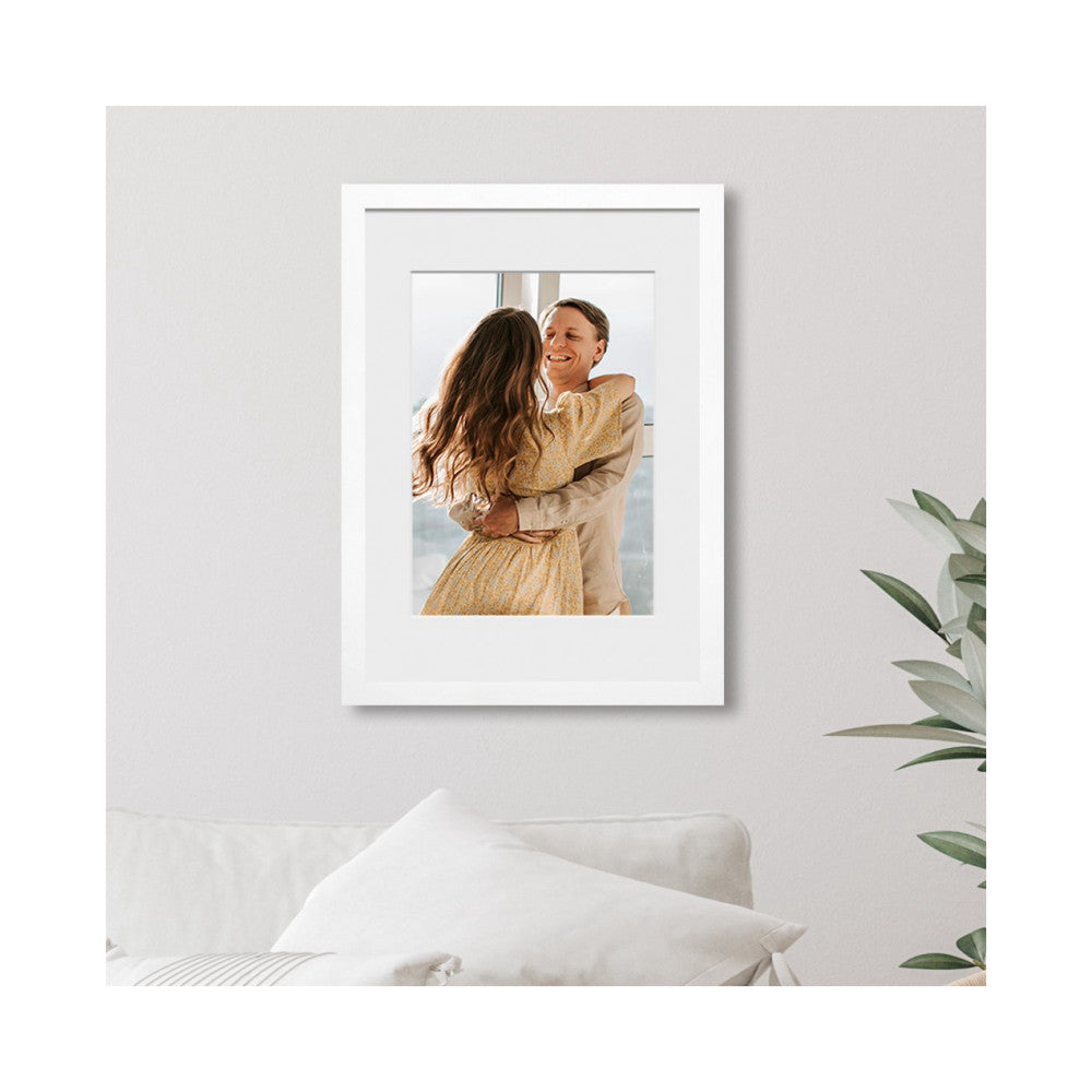 White timber picture frame displaying a portrait photo of a couple embracing, hung on a light wall above a white sofa with greenery visible to the right.
