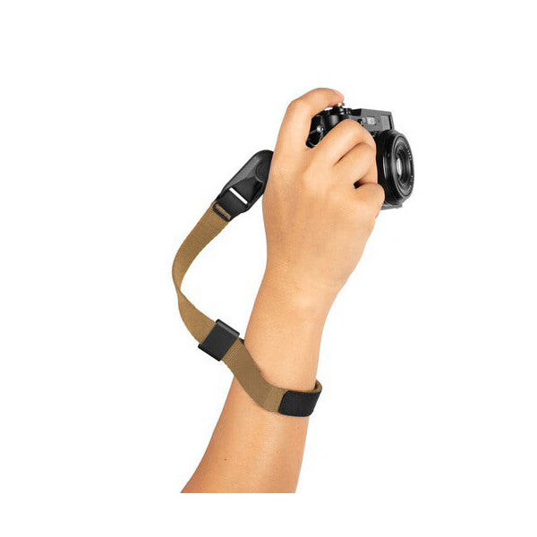 Hand holding a compact camera with a tan and black wrist strap wrapped around the wrist, shown against a white background.