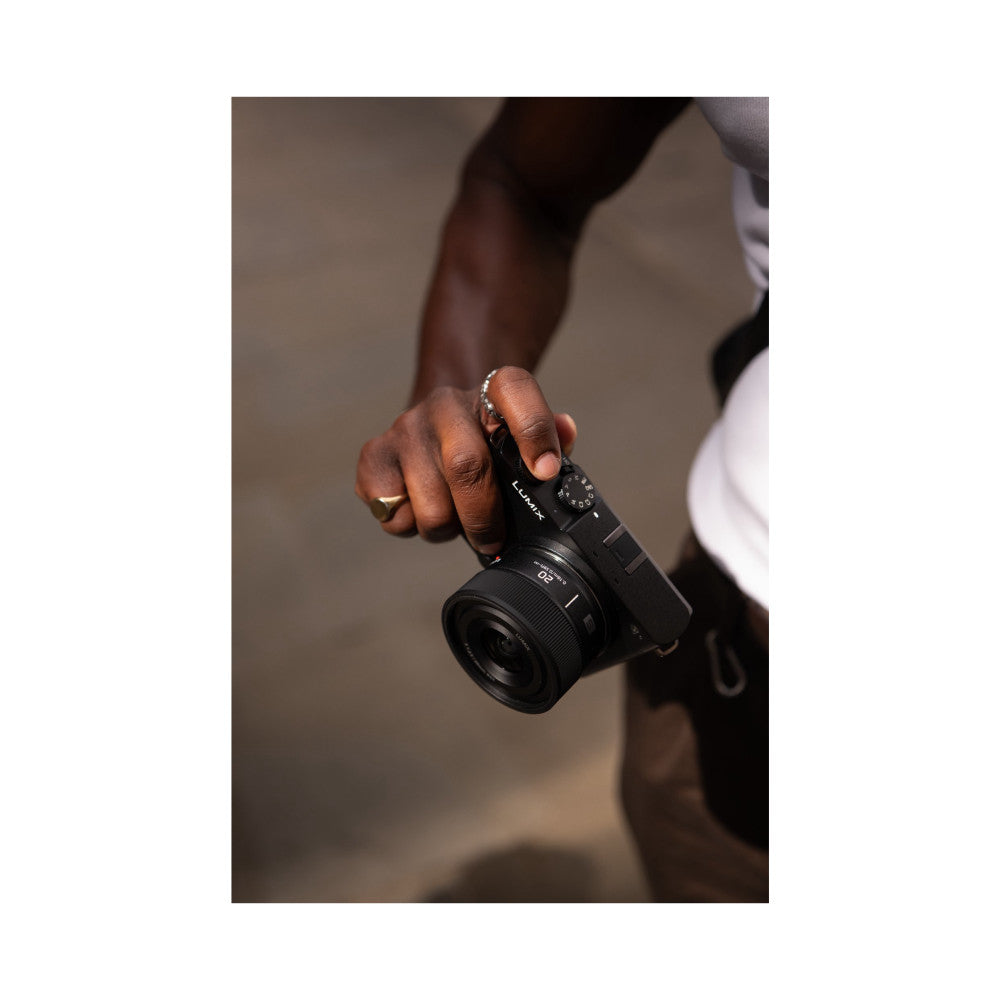 Person holding Panasonic Lumix S camera with 20mm lens, displaying front view of black lens and camera body with visible controls and branding.