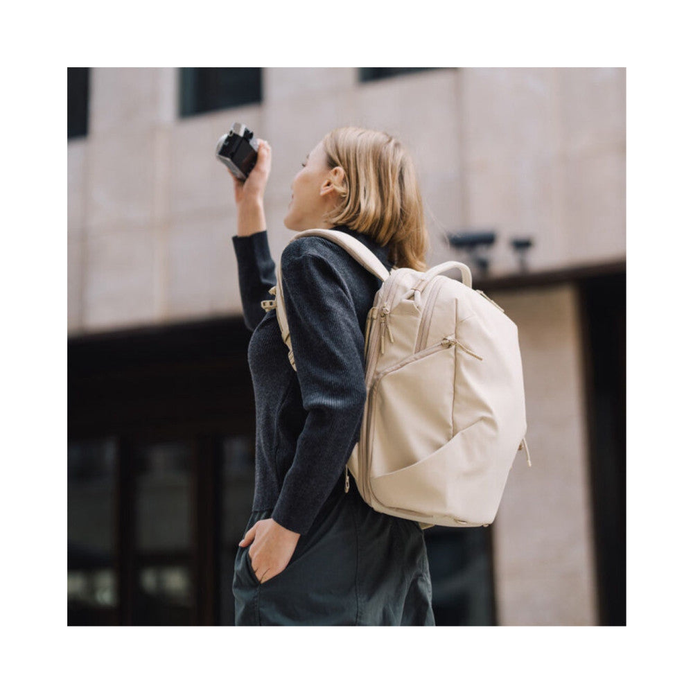 Woman wearing cream backpack shown from rear angle, holding compact camera overhead while looking up at urban building backdrop.