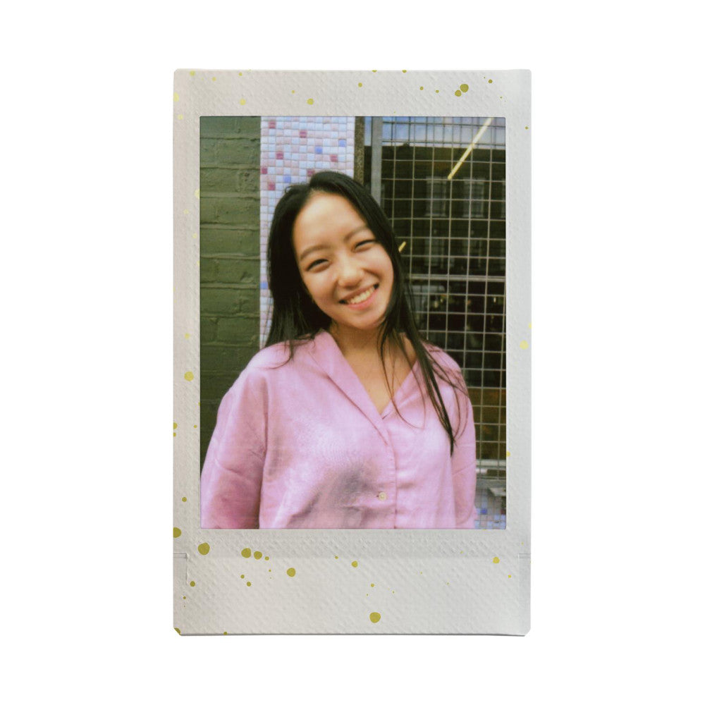 Instant film photo showing a smiling young woman in a pink shirt against an urban background with green brick and metal fence, with gold glitter accents on white border.