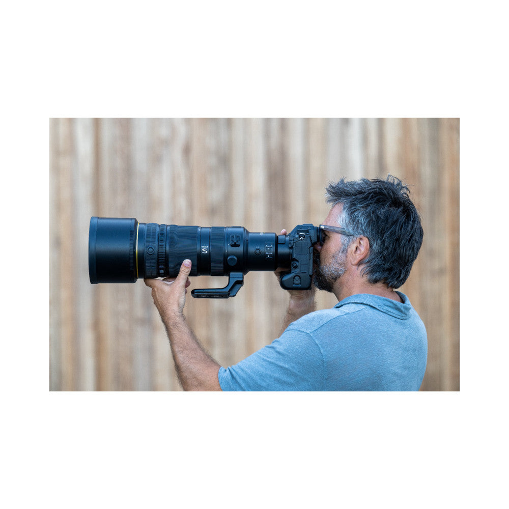 Man wearing glasses using a Nikon Z mirrorless camera with a long black telephoto lens against a blurred beige background.