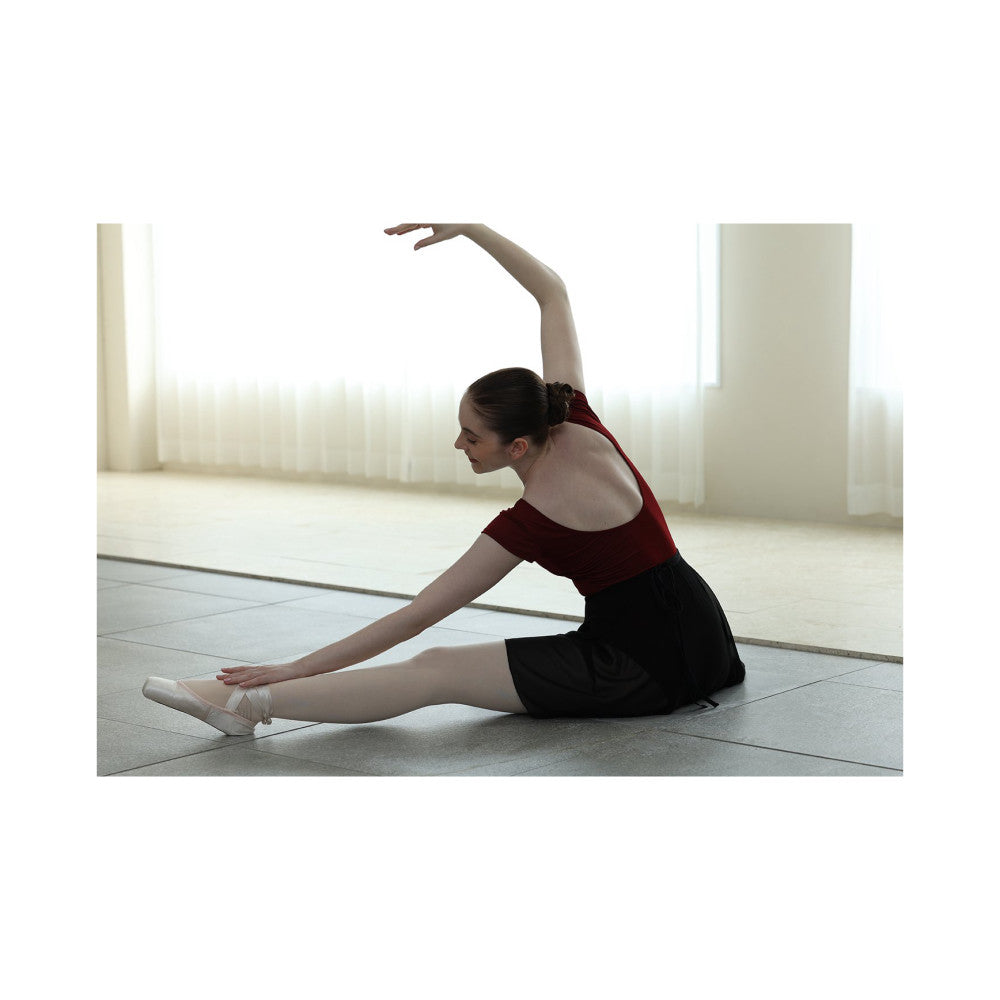 Young ballet dancer in red top and black skirt performing a side stretch on studio floor in pointe shoes.