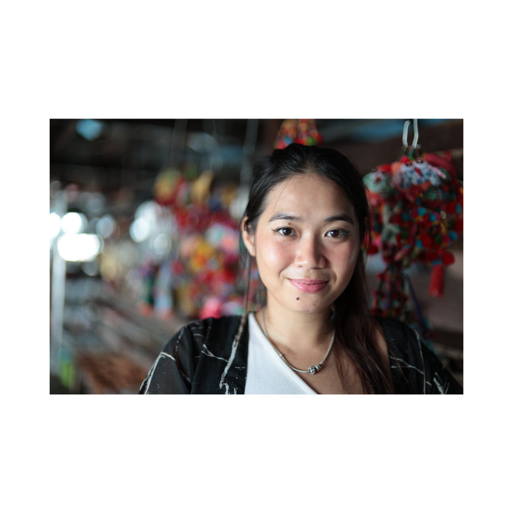 Woman with long dark hair smiling at camera in front of blurred colorful decorations, wearing black jacket and white top.