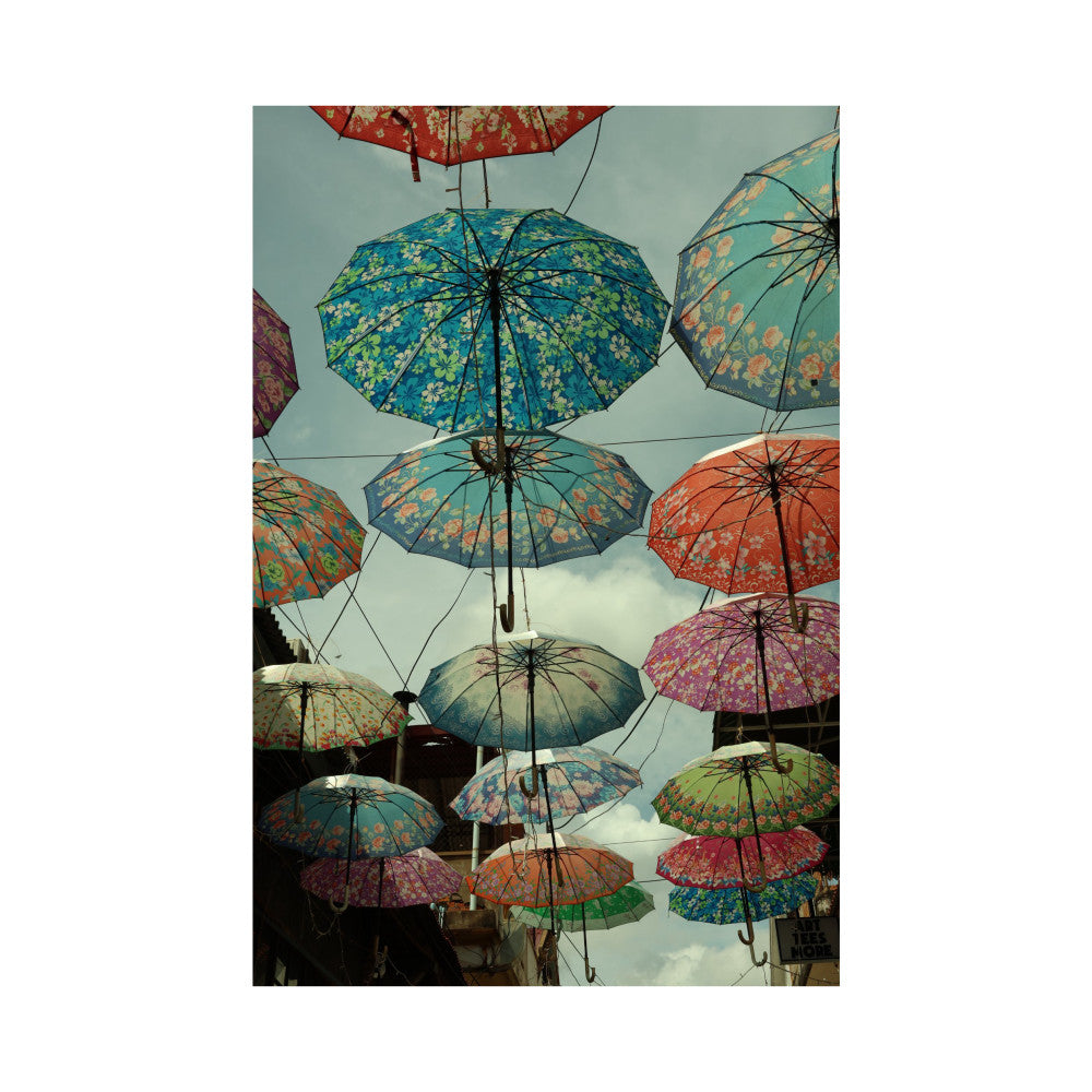 Colorful decorative umbrellas suspended against a cloudy sky, displaying floral patterns in blue, red, green, and orange tones.