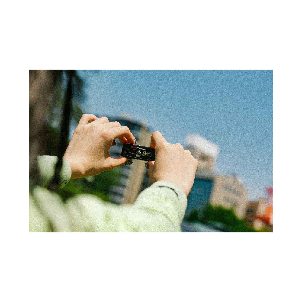 Hands holding a black Kodak Charmera Gold miniature camera keychain, photographed outdoors with blurred cityscape background.