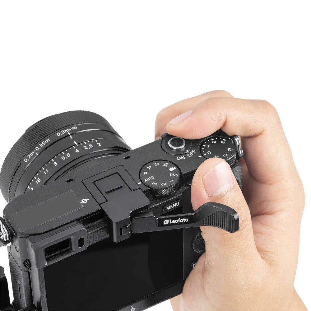 Hand gripping a black camera thumb rest mounted on the right side of a compact camera body with lens attached.