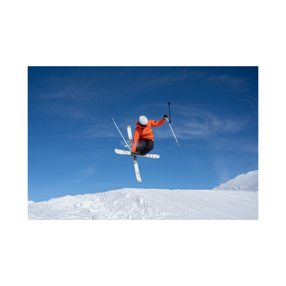 Skier in orange jacket performing aerial trick against blue sky, shot from below showing dynamic action