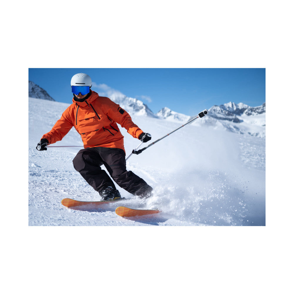 Skier in orange jacket and white helmet descending snowy mountain slope with action camera mounted on pole