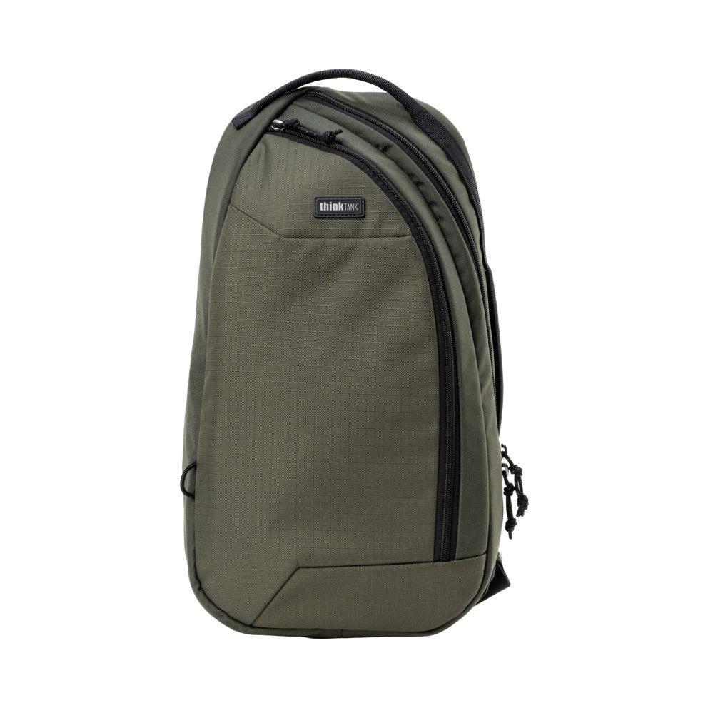 Front view of olive green sling bag with black zippers, padded straps, and thinkTANK branding label on white background.