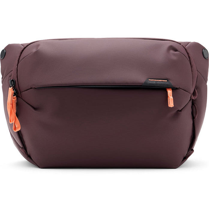 Front view of a burgundy sling bag with orange zipper pulls and rounded corners, displayed against a white background.