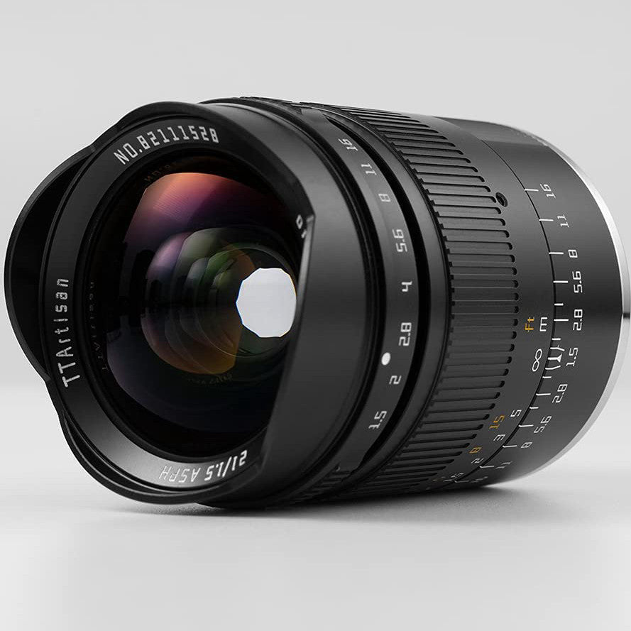 Black TTArtisan 21mm f/1.5 lens shown at a three-quarter angle against a light gray background, displaying the front glass element and focusing ring details.