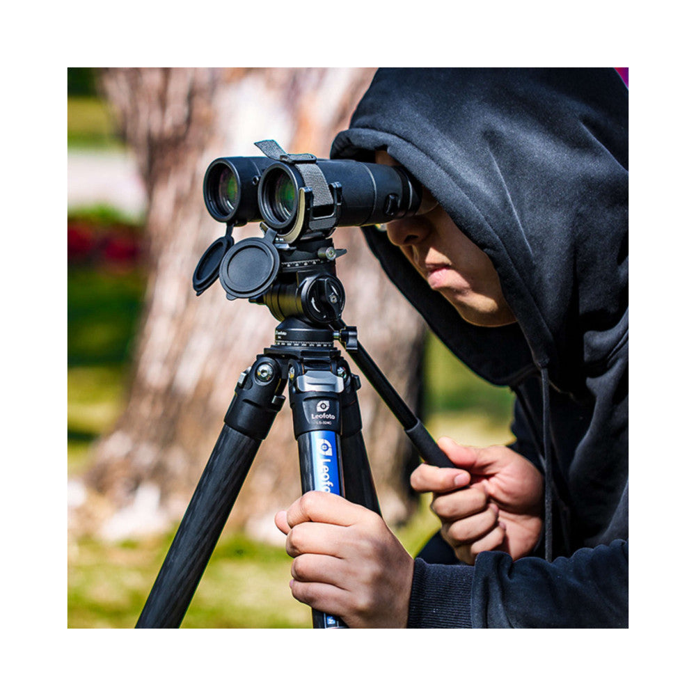 Person in dark hoodie using binoculars mounted on a Leofoto tripod clamp outdoors, demonstrating in-use functionality in a garden setting.