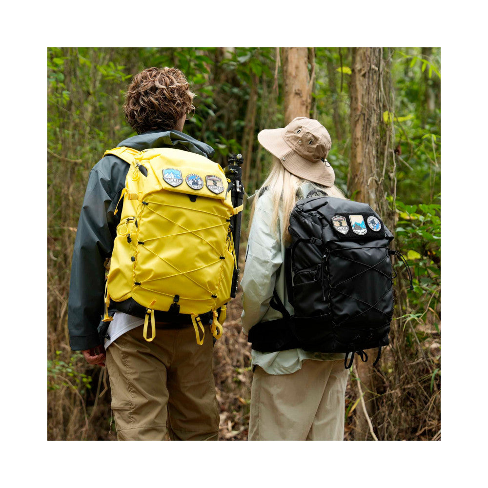 Two hikers wearing camera backpacks on a forest trail, shown from behind, demonstrating the packs in use during outdoor activities.