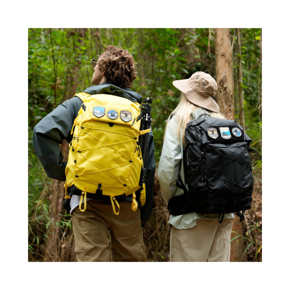 Two hikers wearing large camera backpacks viewed from behind on a forest trail, showing rear pack design and patch details.
