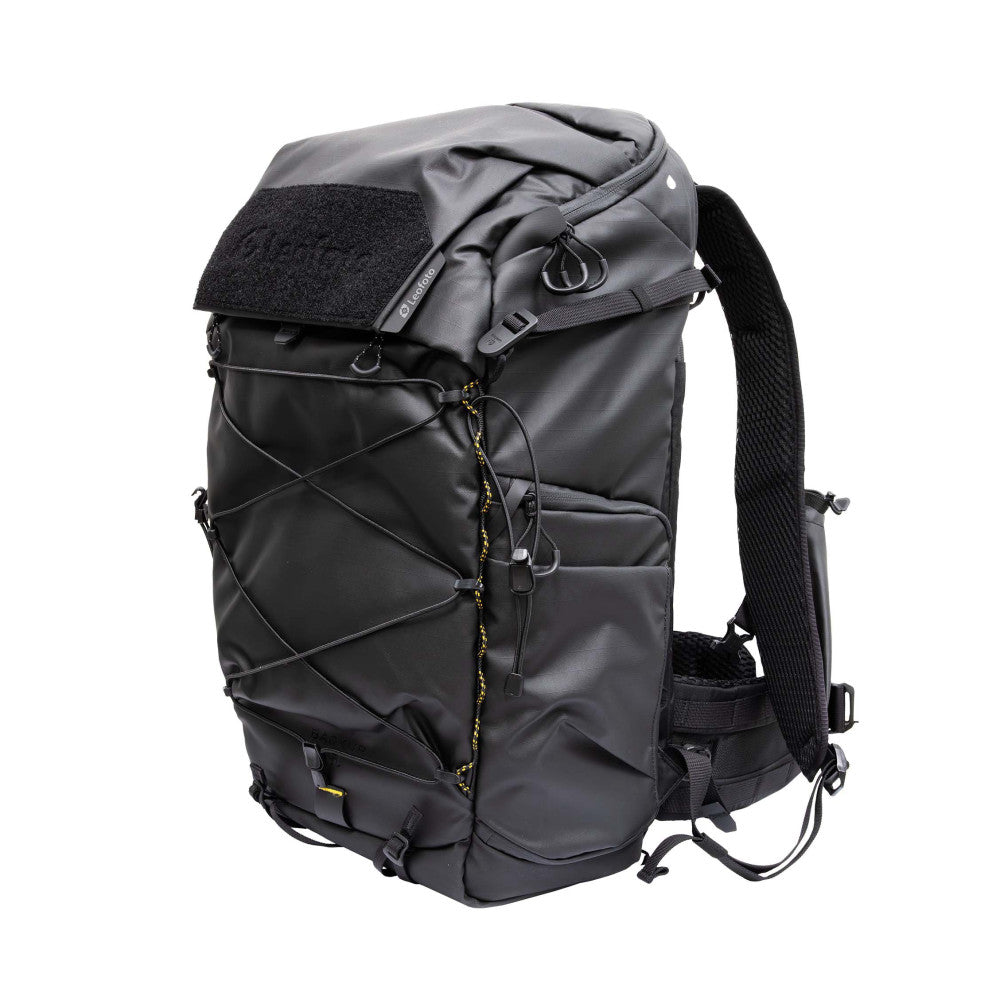 Black camera backpack shown from the rear three-quarter view, displaying padded straps, roll-top closure, and multiple attachment points.