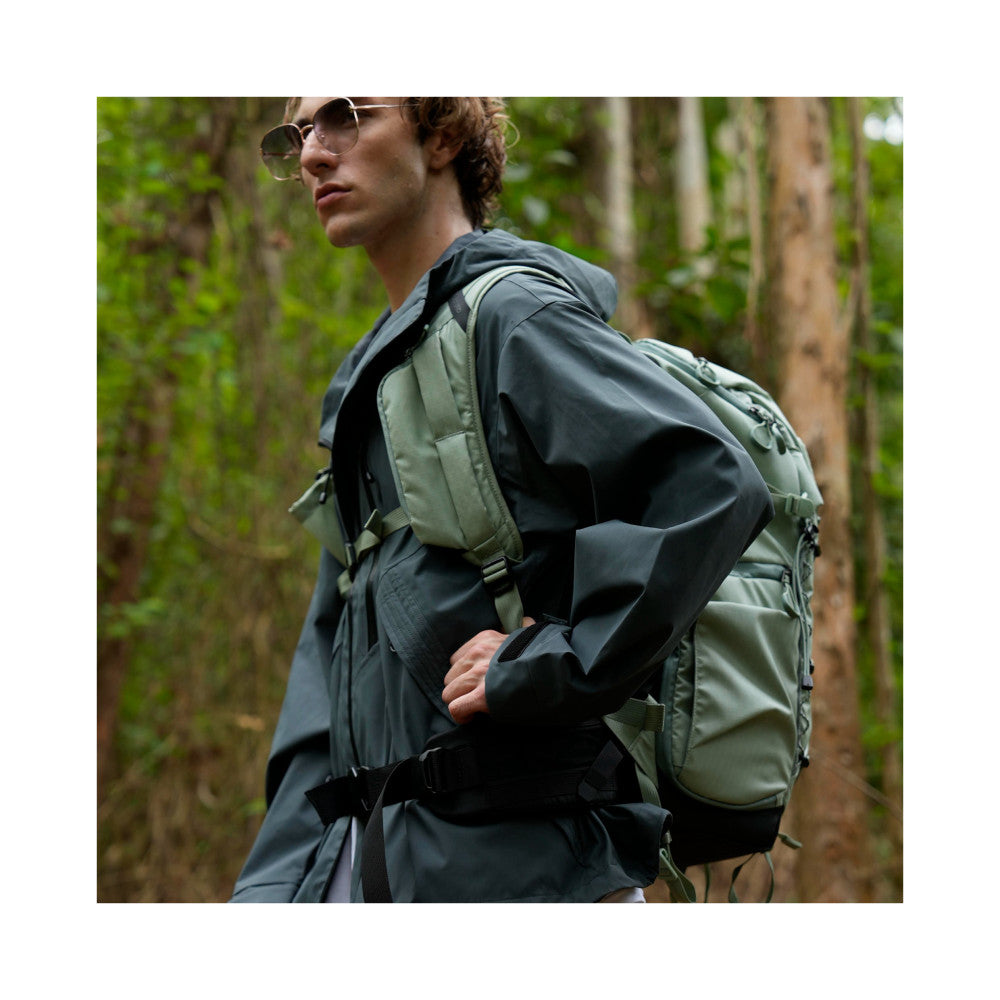 Man wearing green and black camera backpack outdoors in forest, rear three-quarter view showing pack design and fit.
