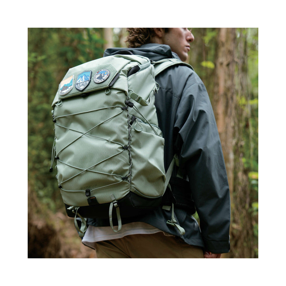 Person wearing green Leofoto camera backpack with patches on shoulder, shown from rear angle in forest setting.
