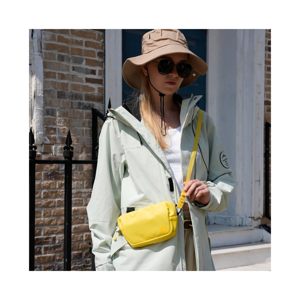 Woman wearing cream jacket and beige bucket hat carrying bright yellow crossbody camera bag, outdoor urban setting, lifestyle product shot.