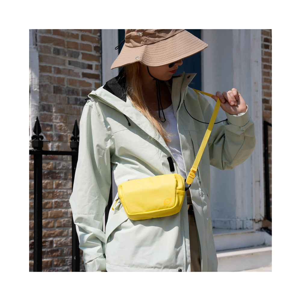 Woman wearing light jacket and bucket hat carrying yellow crossbody camera bag, outdoor street setting, side view.