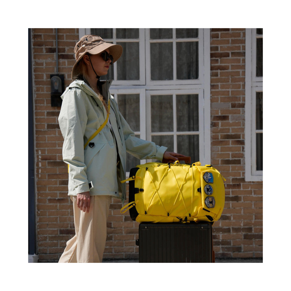 Woman wearing light jacket and cap standing beside yellow camera backpack mounted on black rolling luggage against brick building.