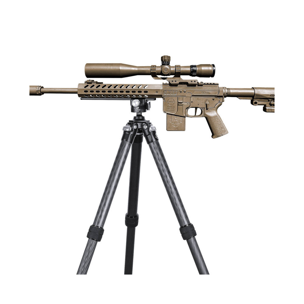 Tactical rifle with tan finish mounted on tripod with gray carbon fiber legs, shown from side angle against white background.