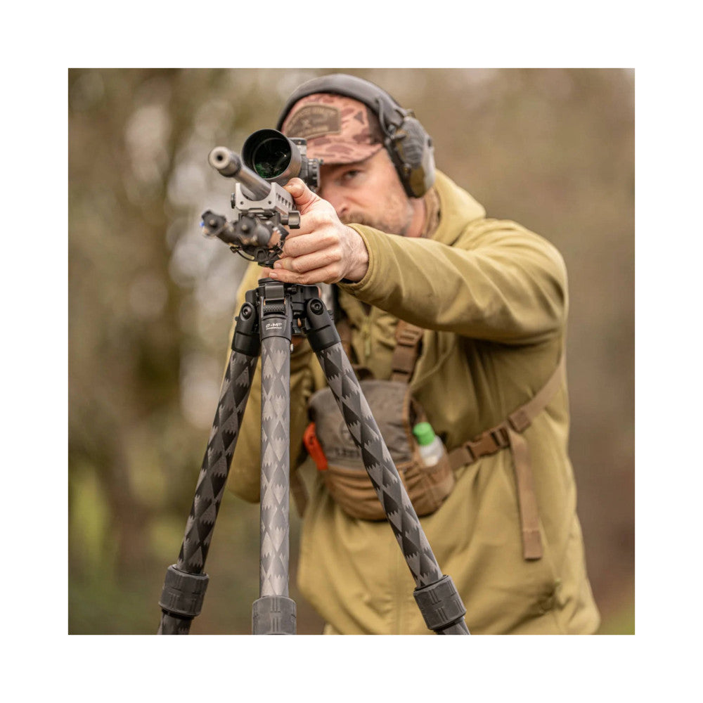 Man aiming rifle with scoped sight mounted on carbon fiber tripod with ball head, demonstrating in-use setup outdoors.