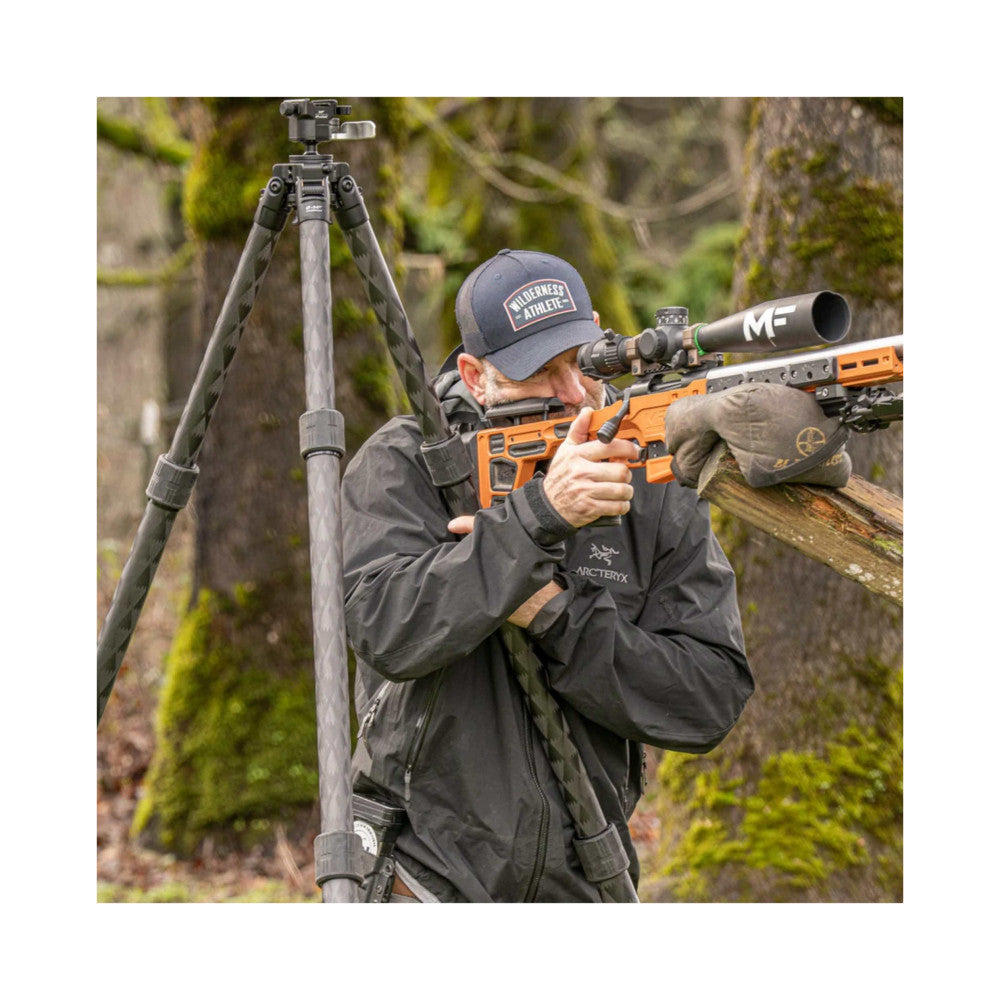 Person aiming rifle with scope mounted on Leofoto carbon tripod in forested setting, demonstrating in-use stability and support.