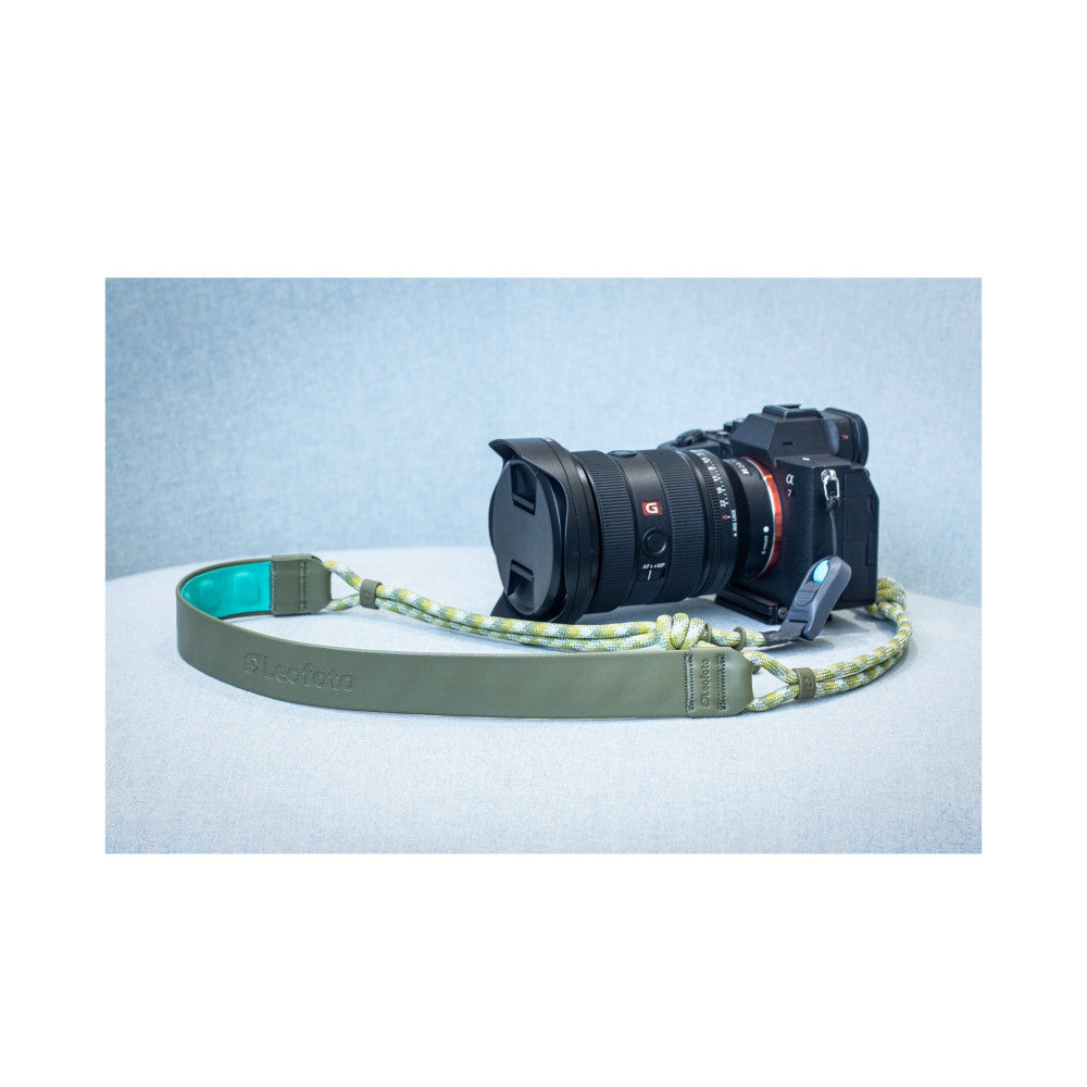 Pine green camera strap with rope detail displayed on white surface next to black mirrorless camera with lens attached.