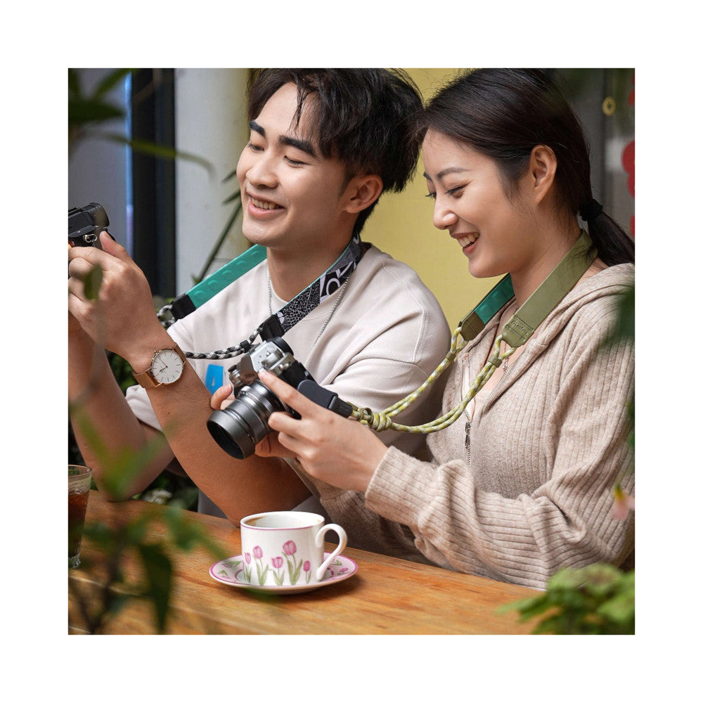 Two people reviewing photos on a camera equipped with a green camera strap, seated at a wooden table with a floral teacup nearby.