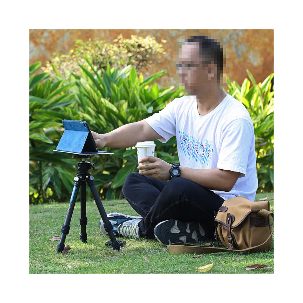 Person outdoors using tripod-mounted tablet tray while holding beverage, demonstrating portable product setup in garden setting.