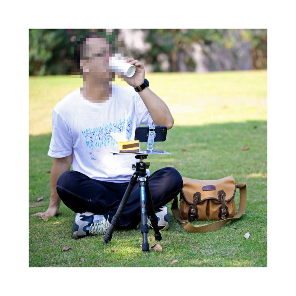 Person demonstrating Leofoto LCH-1 tray mounted on tripod outdoors, holding cup with smartphone and accessories attached to the platform.