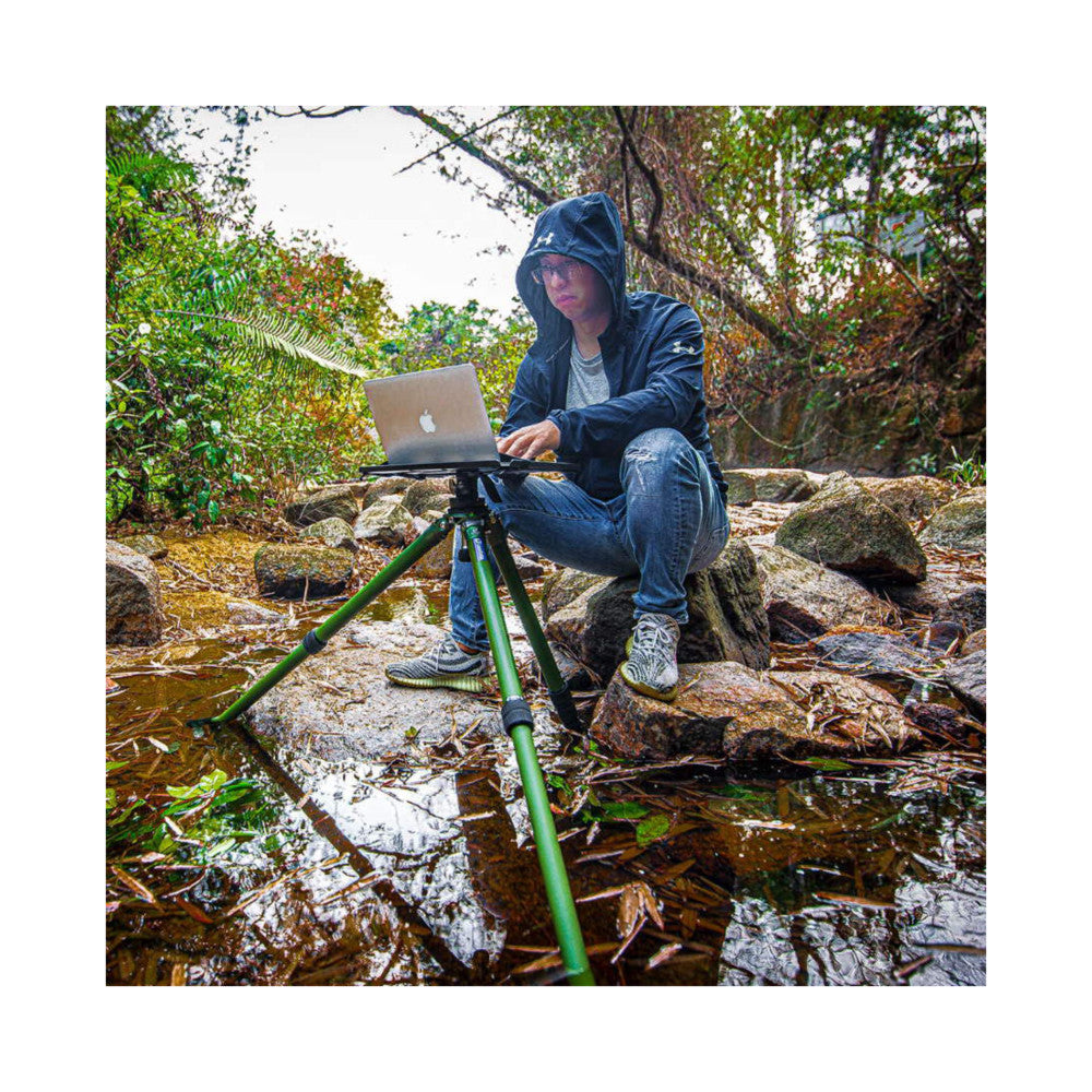 Person using green tripod-mounted laptop tray in outdoor forest stream setting, demonstrating in-use functionality with MacBook.