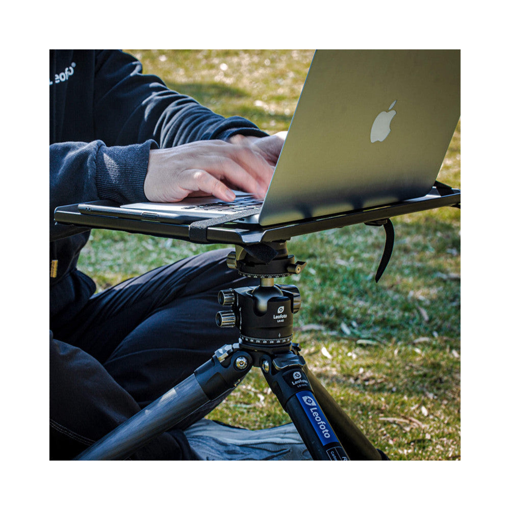 Person using Leofoto tripod with laptop tray holding MacBook outdoors on grass, showing product in-use setup