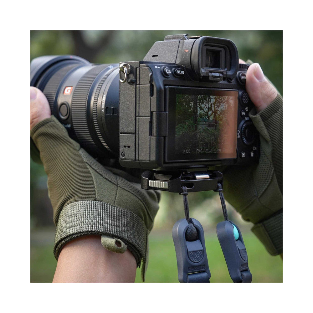 Sony mirrorless camera with lens attached, shown from rear angle held in hands wearing olive gloves, with two black quick release clamps hanging below the camera body.