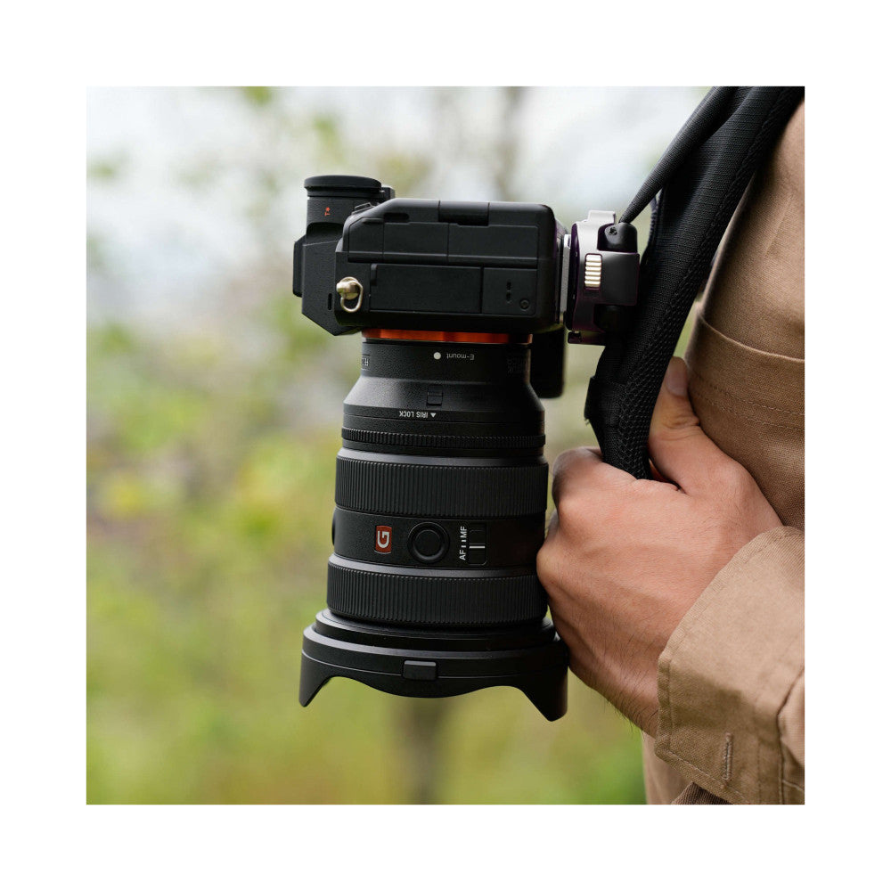 Hand holding a camera lens and tripod head mounted with quick release plate, showing the connection mechanism from a side angle outdoors.