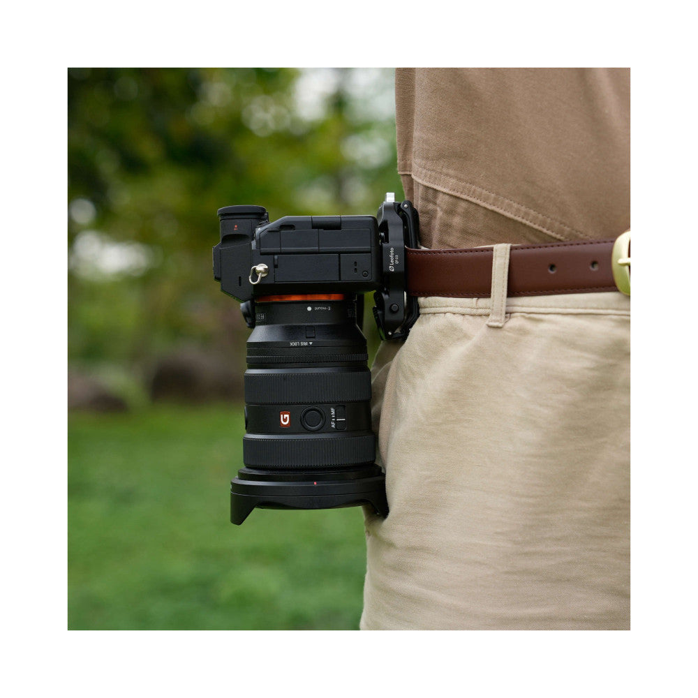 Black camera quick-release mount attached to belt, shown in-use from side angle with lens mounted on outdoor background.