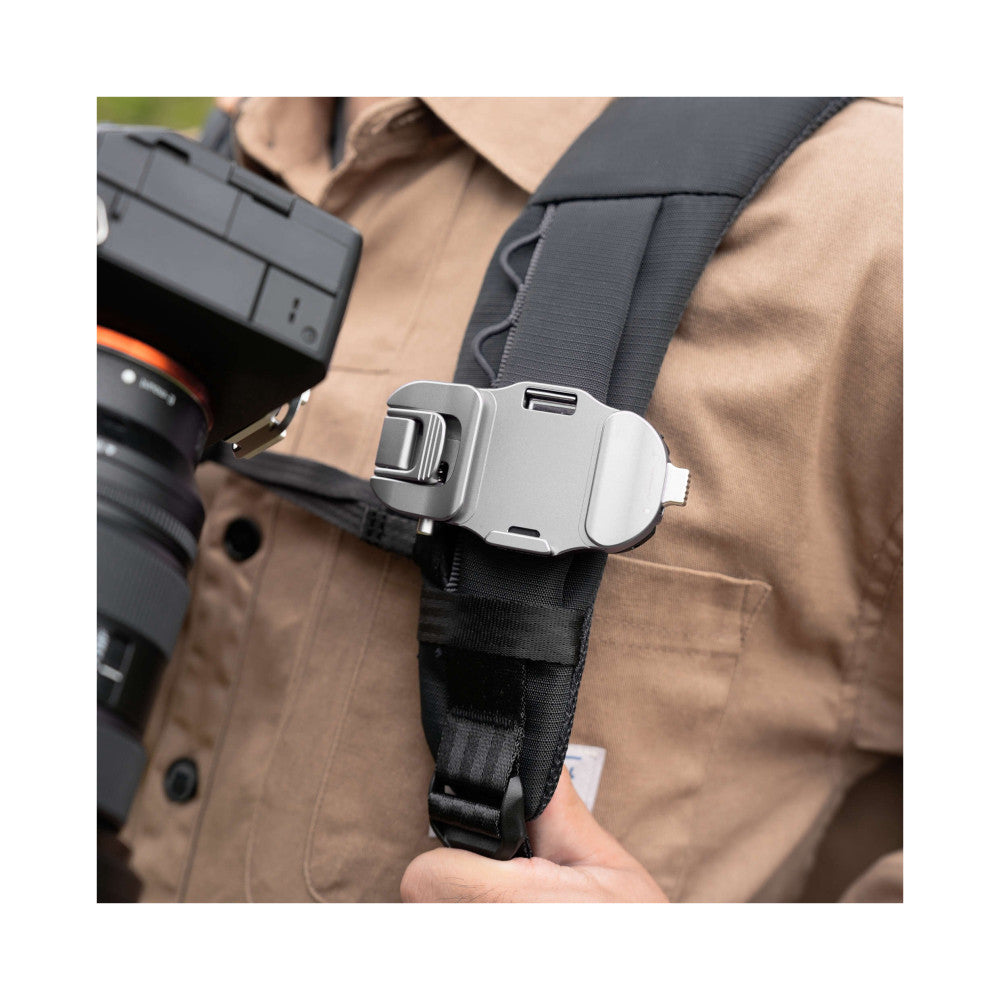 Silver quick release camera mount attached to black strap on tan vest, shown in close-up detail from front angle.