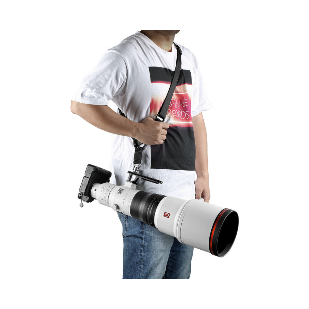 Person wearing white graphic tee carrying professional camera with telephoto lens using black shoulder strap, shown from side angle.