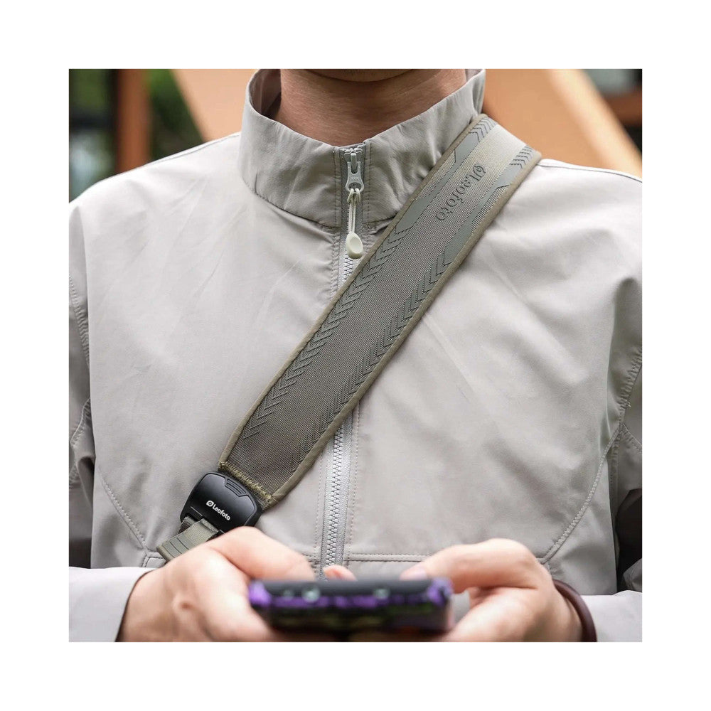 Person wearing light gray jacket with olive green camera strap across chest, showing strap detail and quick-release connector in use.
