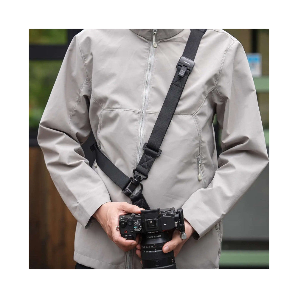 Person wearing light gray jacket with black camera strap across chest, holding professional video camera with quick-detach strap system in use.