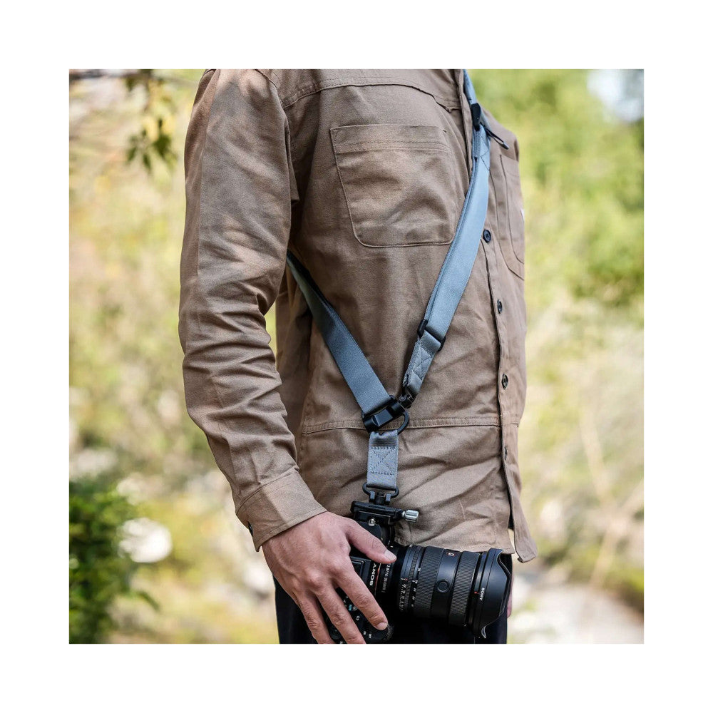 Person wearing brown jacket holding DSLR camera with gray camera strap attached, shown in-use from side angle outdoors.