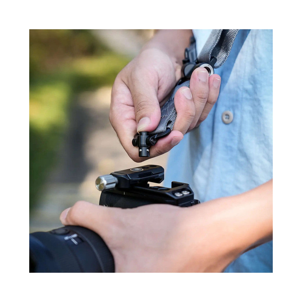 Hands demonstrating quick-detach camera strap attachment system with gray strap and black metal connector on camera equipment outdoors.
