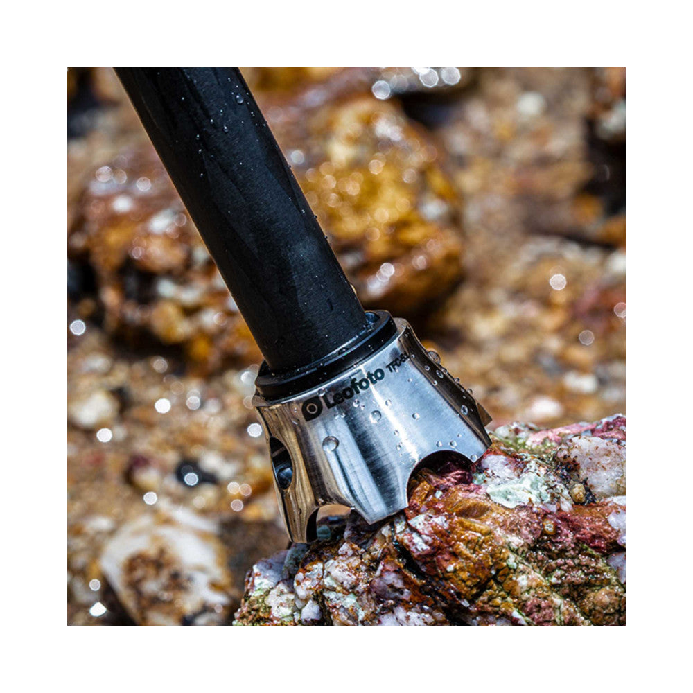 Close-up of silver tripod claw attachment mounted on black leg, gripping colorful coral rock underwater with water droplets visible.