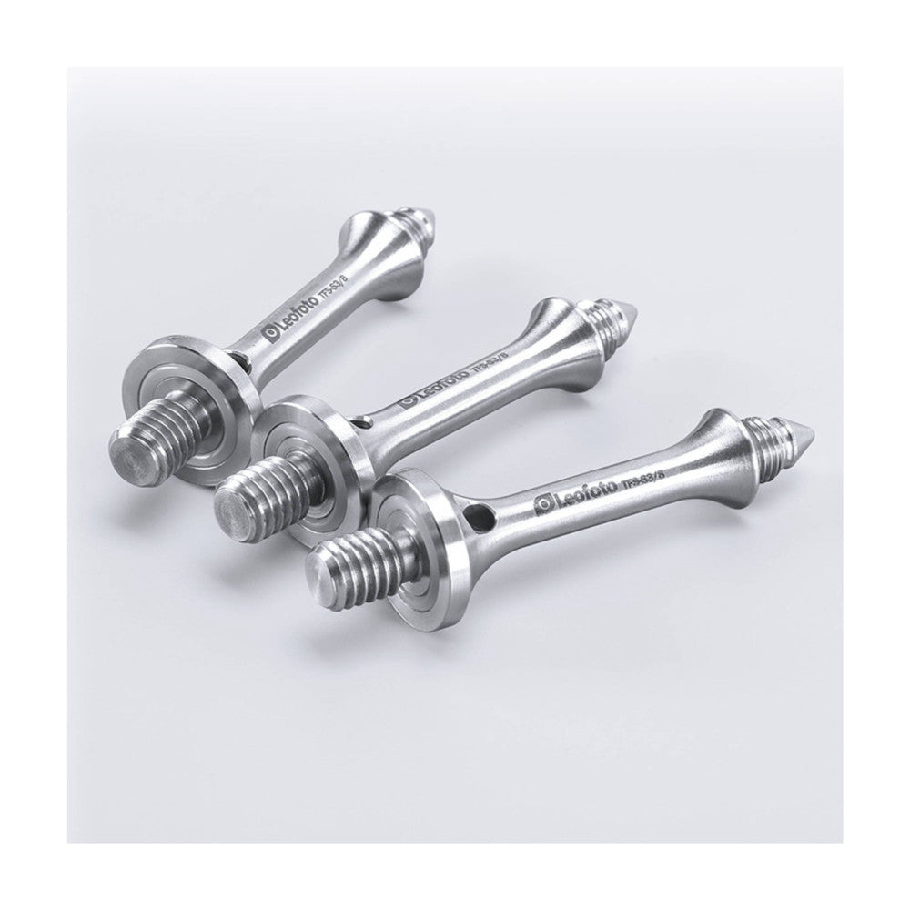 Three polished metal tripod spikes with threaded bases arranged at angles against a light gray background, showing their tapered points and mounting hardware.