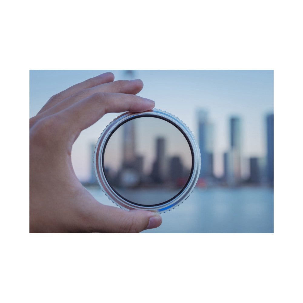 Hand holding a circular camera filter with blue-tinted metal ring, showing the clear glass center with city skyline blurred in background.