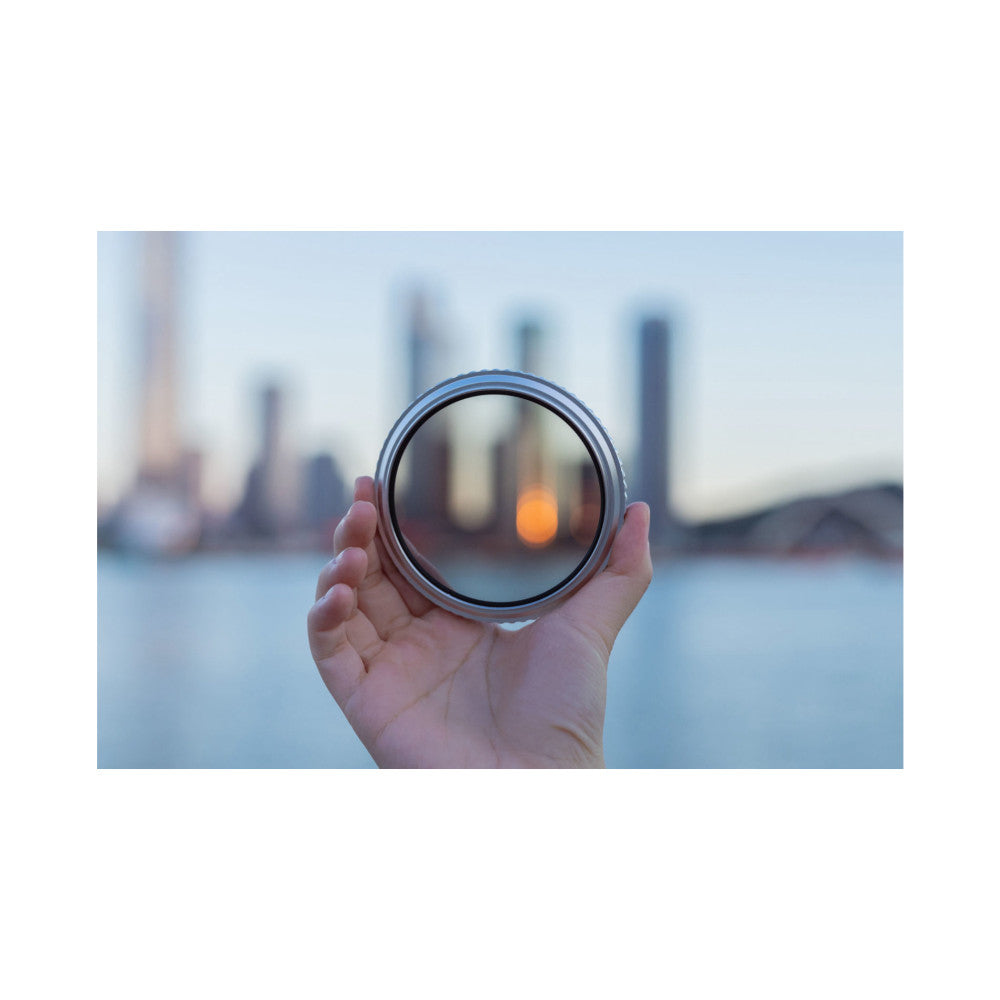 Hand holding circular camera filter against blurred cityscape, showing filter's clarity and light transmission through glass element.