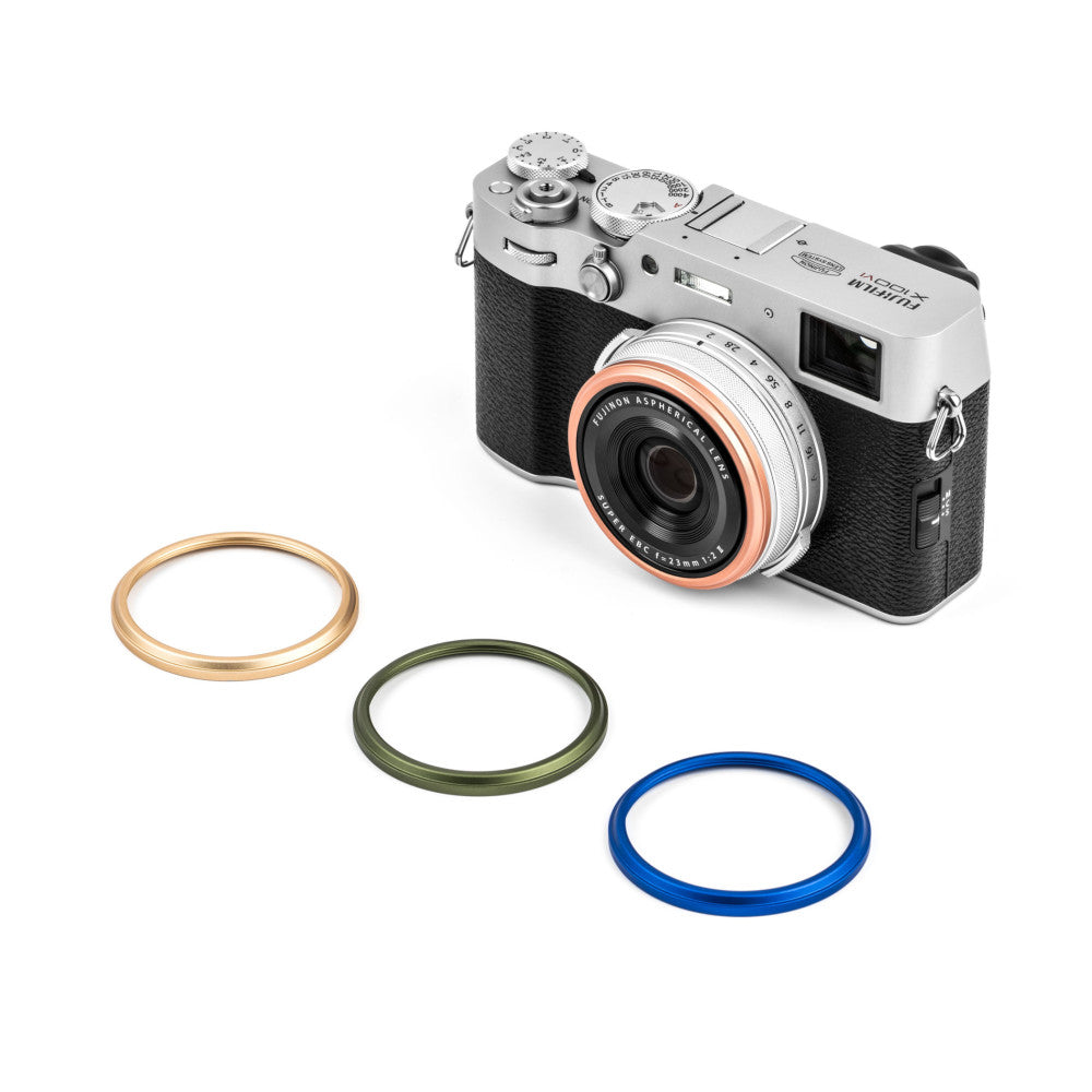 Fujifilm X100 camera with four colored lens rings displayed separately below: gold, rose gold, green, and blue.